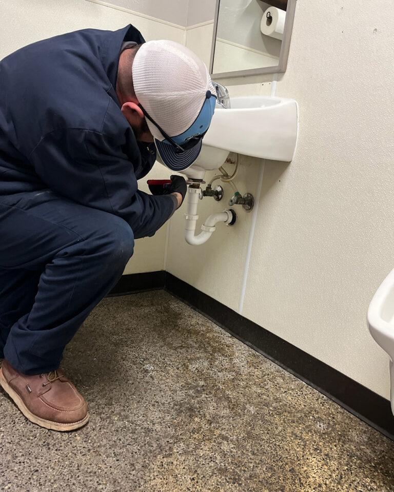 A person in workwear crouches under a bathroom sink, inspects the plumbing, and checks the faucets.