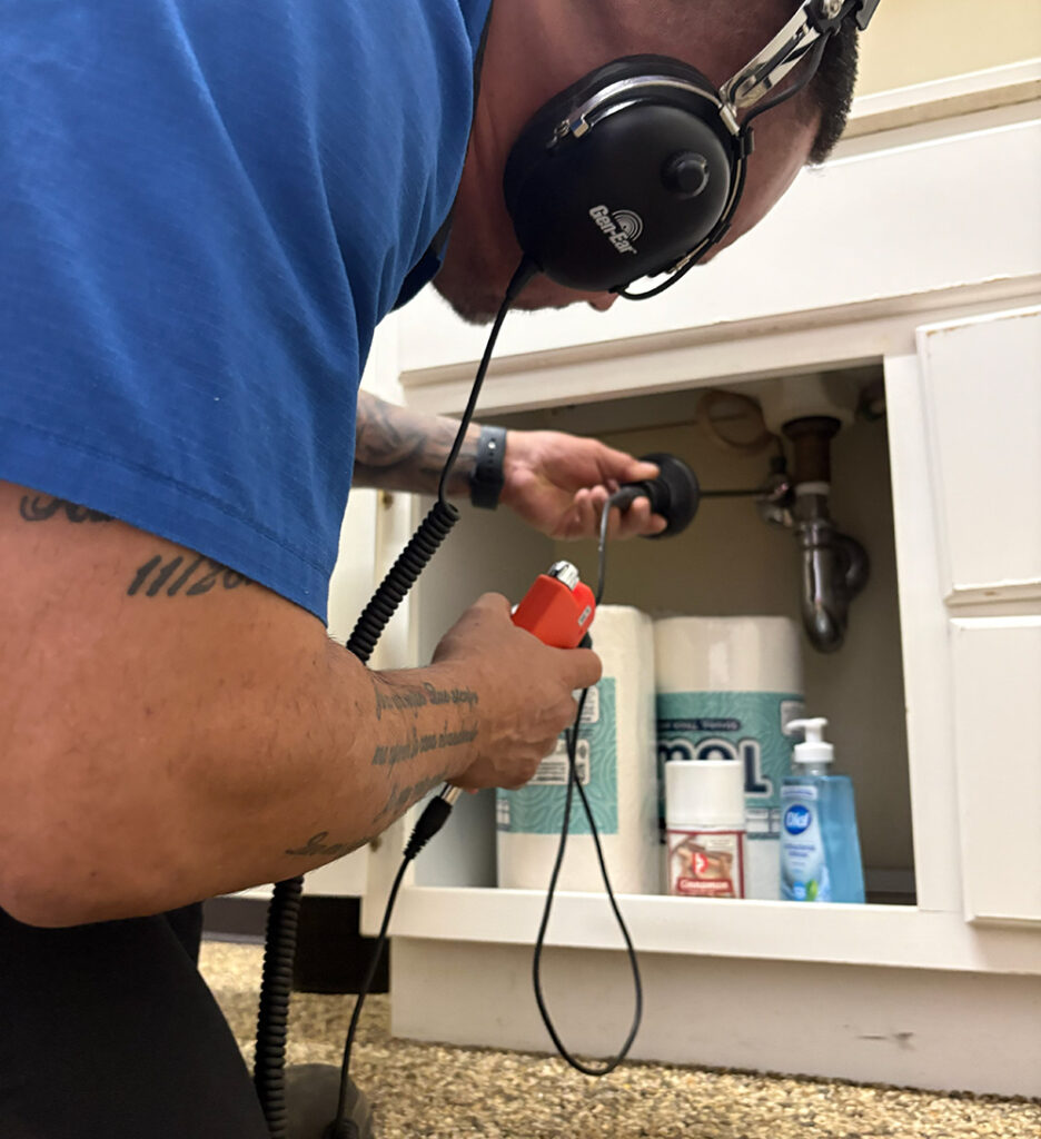 Wearing headphones, a person inspects plumbing under a bathroom sink with a leak detector, near stored toilet paper and soap.
