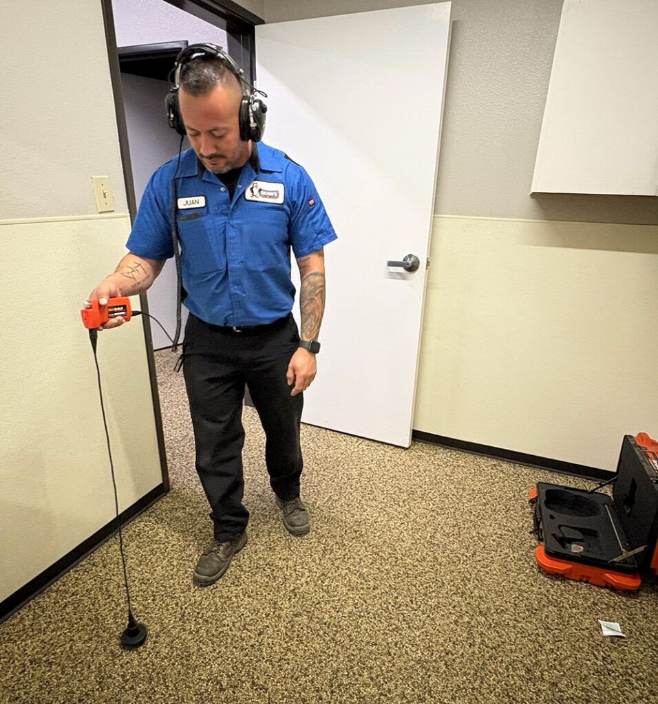 A man in work attire inspects the carpeted office floor for leaks using electronic equipment, with a black case and tools nearby.