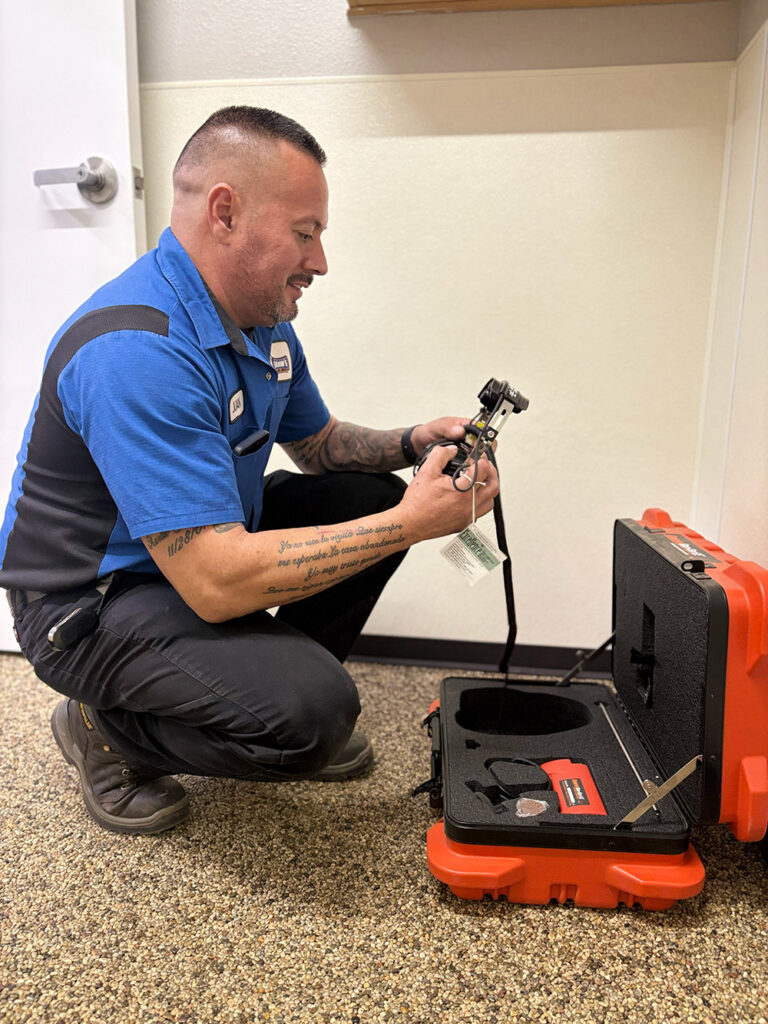 A man in a blue work shirt kneels on the floor and inspects a leak detection tool from an open orange foam-padded hard case.