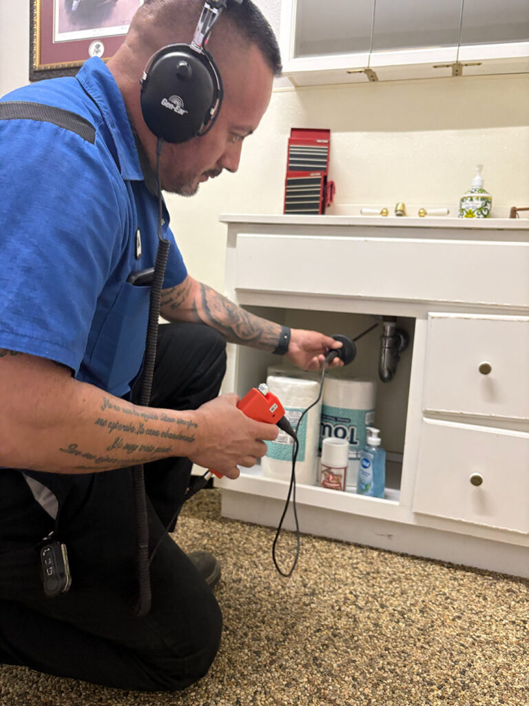 Wearing headphones, a plumber checks for leaks with an electronic device under a bathroom sink; toiletries fill the cabinet below.