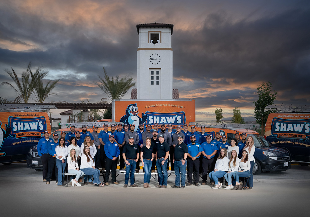 Shaw’s Home Comfort employees stand in front of company vans and a clock tower building beneath a dramatic sky.