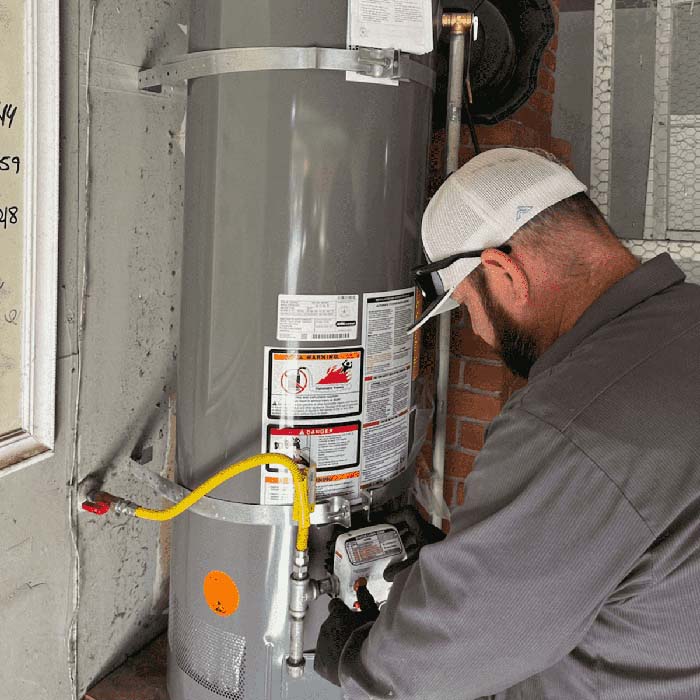 A man in work clothes and a cap inspects or repairs a gray water heater by a brick wall to keep it running efficiently.