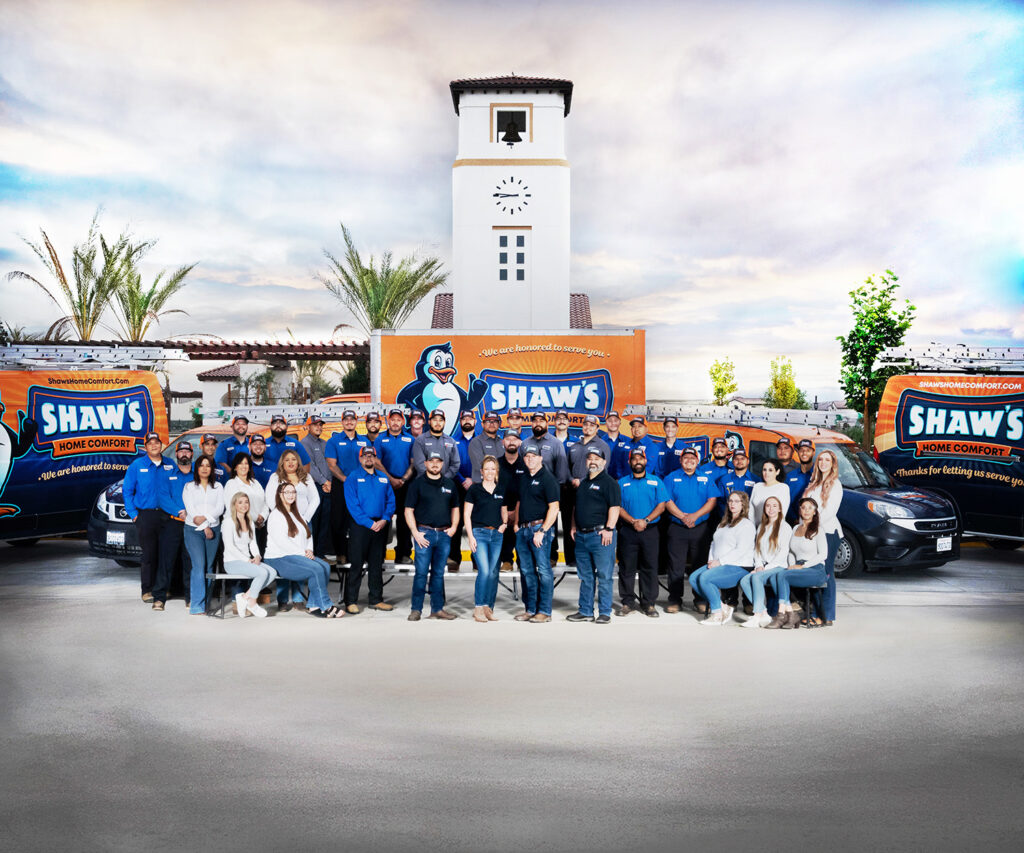 Shaw's Home Comfort employees pose in front of two company vans and a clock tower building, wearing casual and branded uniforms.