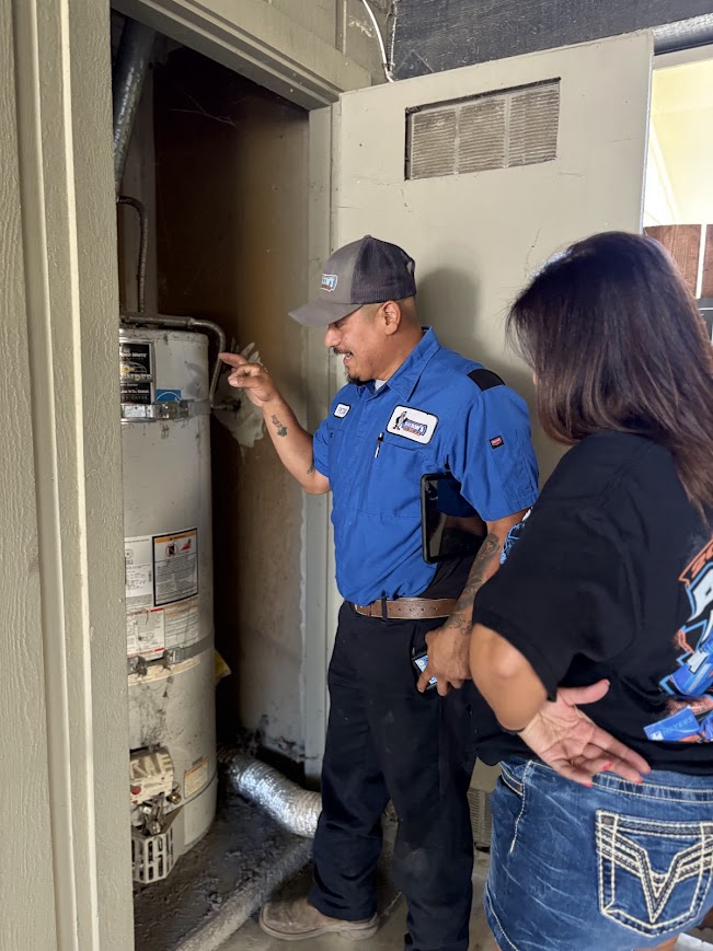 A technician in a blue uniform explains the water heater to a woman in a black shirt inside a utility closet.